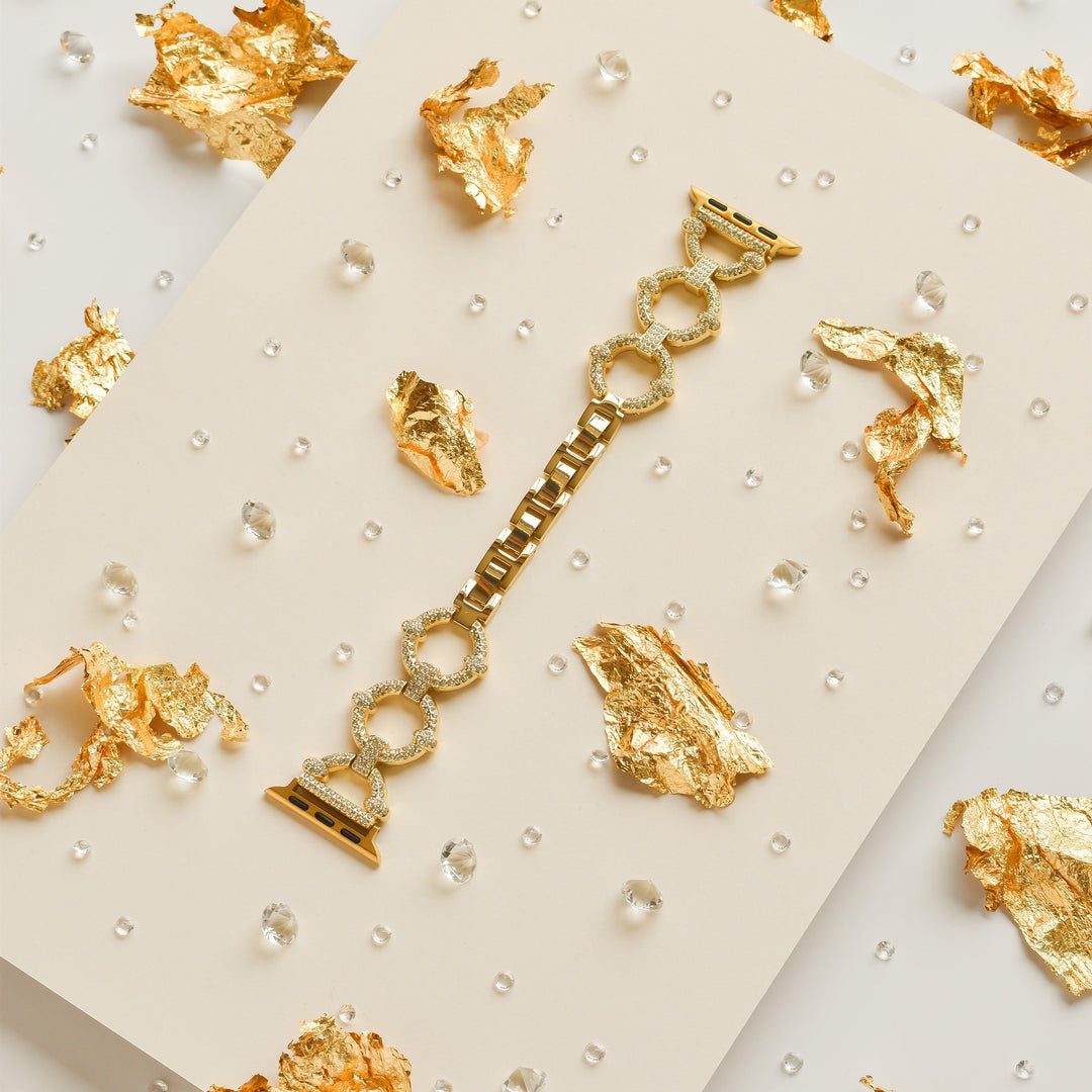 The Diamante Bracelet Apple Watch Strap by Coconut Lane is showcased on a cream-colored surface, adorned with scattered pieces of gold foil and small clear beads for an elegant and luxurious ambiance. Featuring large circular links in a gold finish, this strap fits various Apple Watch models and adds a touch of sophistication.