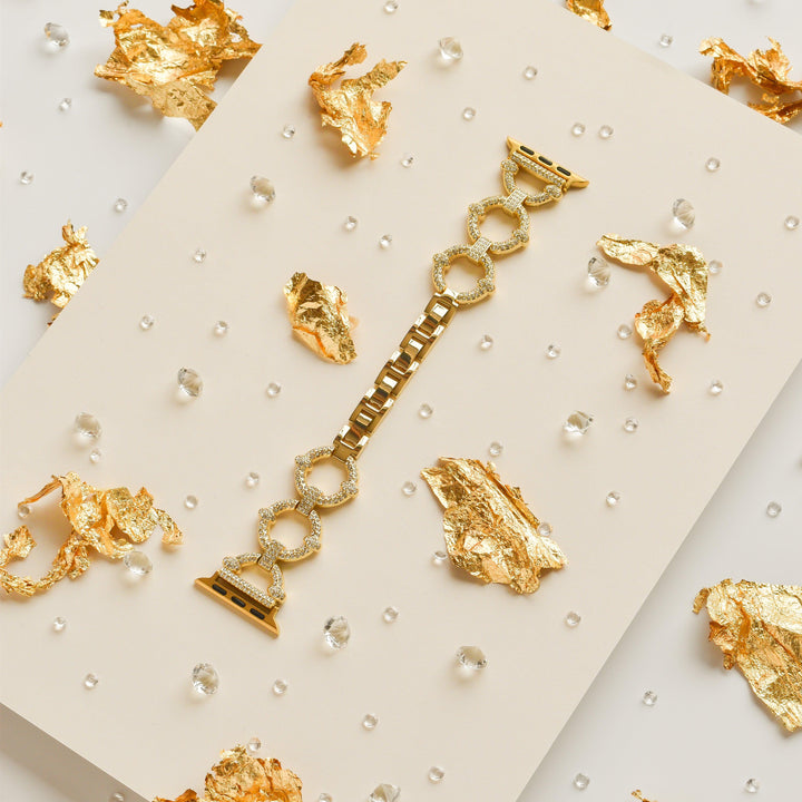 The Diamante Bracelet Apple Watch Strap by Coconut Lane is showcased on a cream-colored surface, adorned with scattered pieces of gold foil and small clear beads for an elegant and luxurious ambiance. Featuring large circular links in a gold finish, this strap fits various Apple Watch models and adds a touch of sophistication.