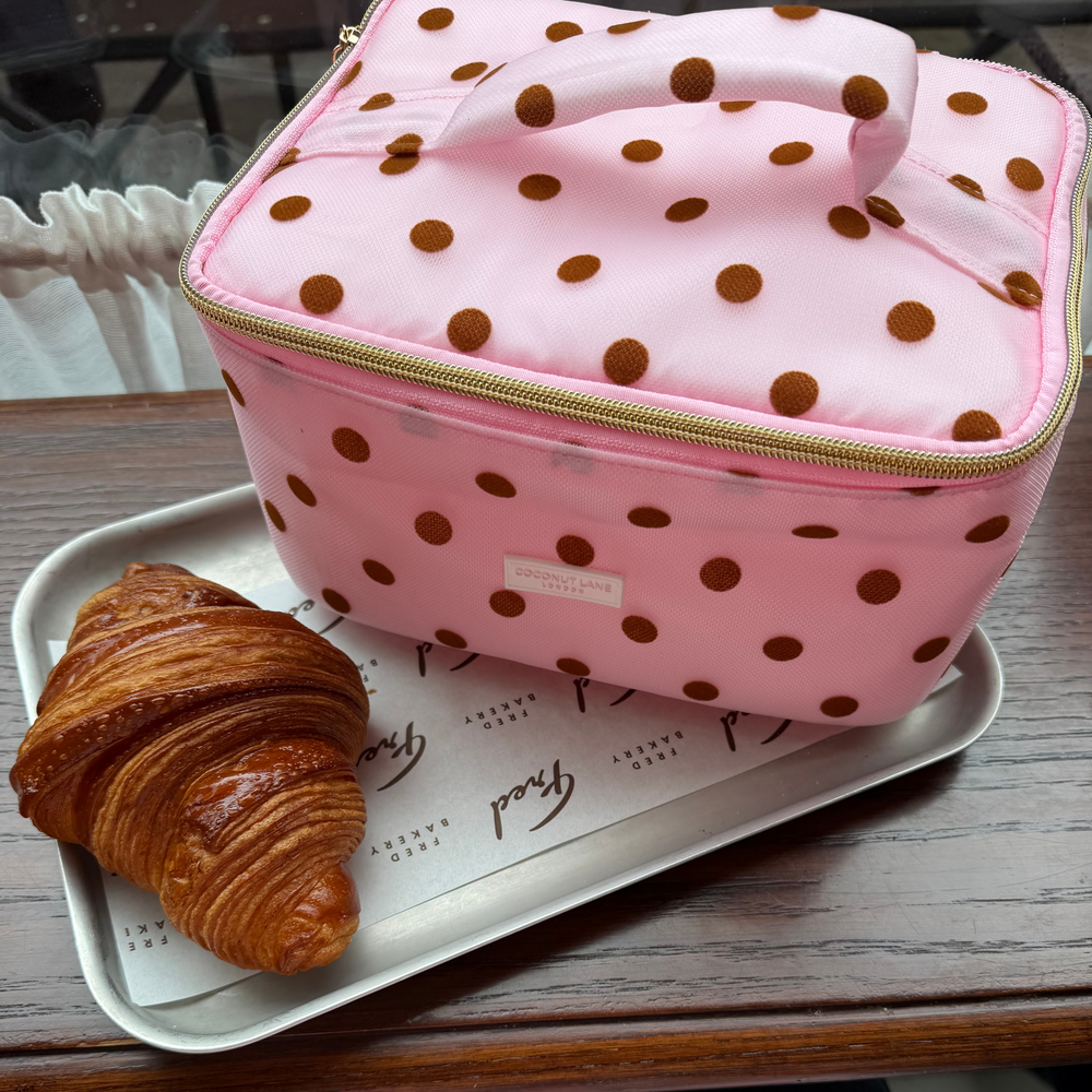 Flat lay photo in a cafe of the Dot Society pink and brown vanity case. The vanity case is baby pink mesh type fabric, with chocolate brown polka dots. 
