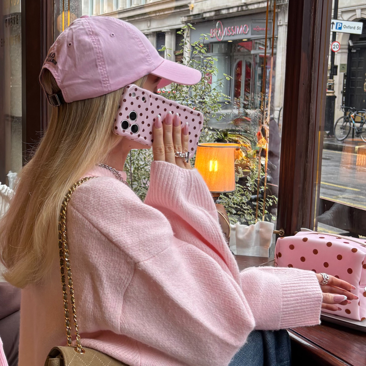 Woman wearing baby pink jumper and cap, holding an iphone up to her ear. The iphone has a wavy baby pink phone case with brown polka dots.