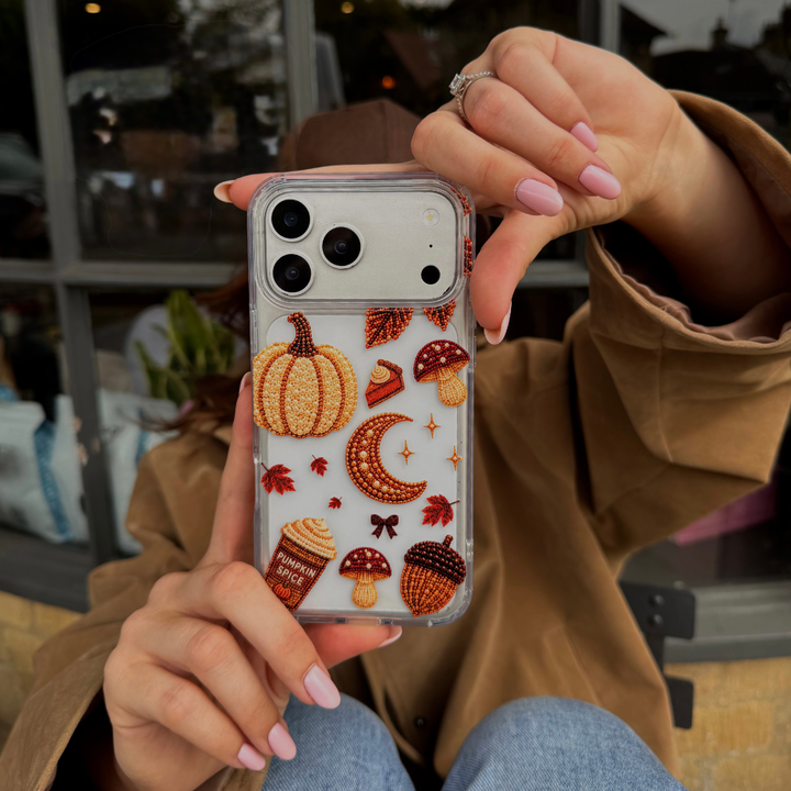 A person holds a clear phone case decorated with autumn-themed beaded effect designs including pumpkins, acorns, mushrooms, leaves, and a pumpkin spice coffee cup, shown in a lifestyle cafe setting.