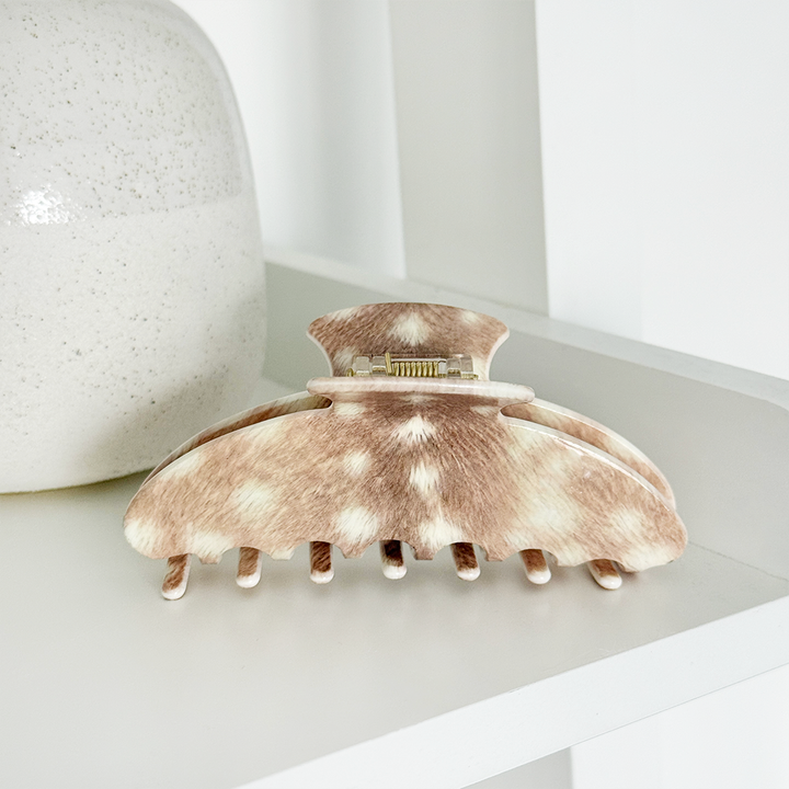 Hair claw clip with a brown and cream mottled animal-print pattern, resting on a white shelf next to a textured ceramic vase.