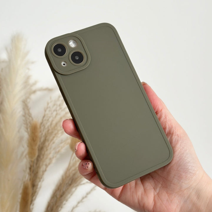 A hand holding a smartphone with a Coconut Lane NAKD Phone Case - Khaki. The phone features dual cameras and an LED flash, providing full camera protection. In the background, minimalism is showcased by dried pampas grass stalks against a light-colored wall.