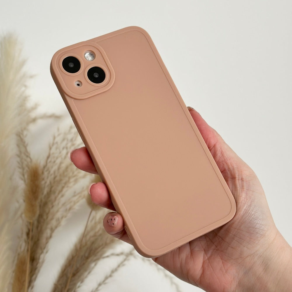 A hand holding a smartphone with a light peach Coconut Lane NAKD Phone Case against a neutral background. Dried pampas grass is visible in the background. The phone case, one of our pastel favourites, has a smooth, matte finish with cutouts for the camera lenses and flash.