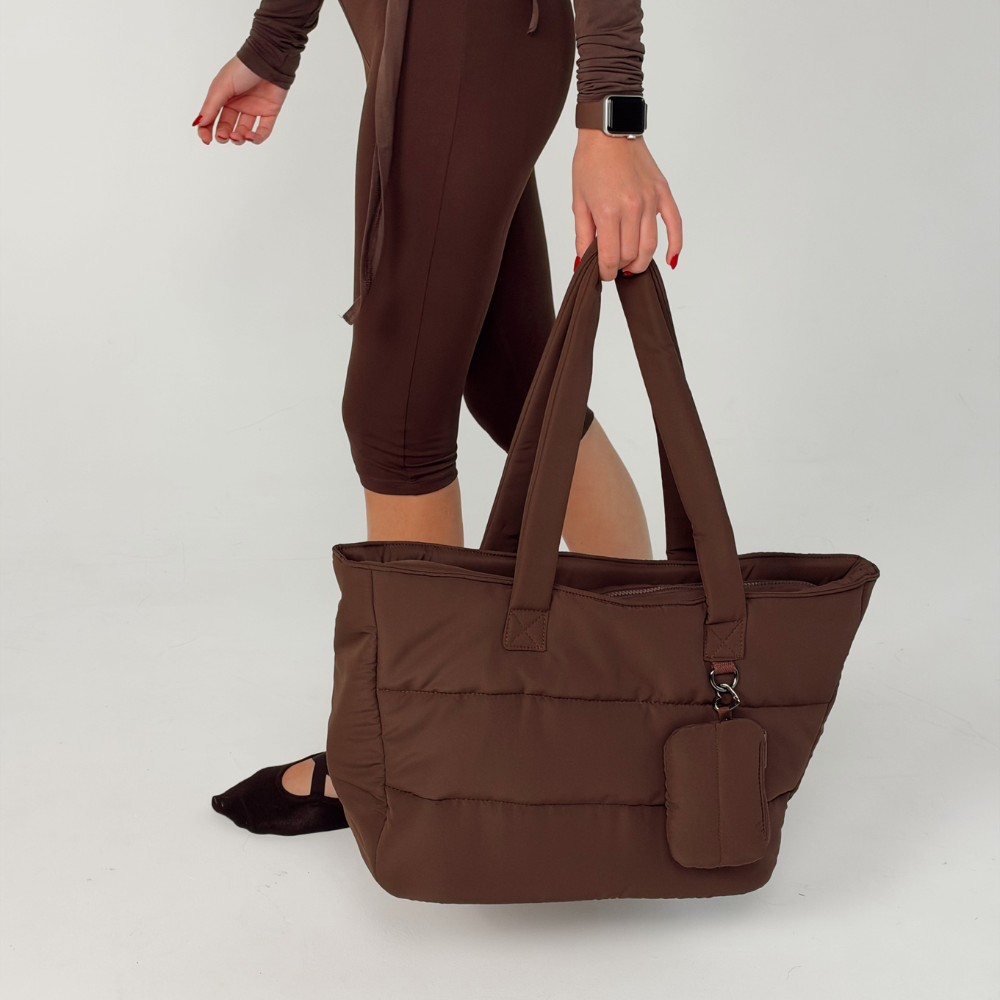Woman in a brown pilates outfit, holding the Coconut Lane Main Carry Choc Puffer bag with the matching Chocolate brown mini keyring bag with zip fastening, silver metal heart shaped clip attached to the outside D ring. On her wrist is the Coconut Lane NAKD apple watch strap in chocolate brown.
