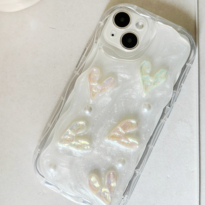 A close-up image of an iPhone with a Coconut Lane Pearly Hearts Phone Case - Clear. The case, clear with a pearl white shimmer effect, is adorned with raised iridescent heart shapes and small pearl-like beads. Extra protective, the phone is placed on a light gray surface.