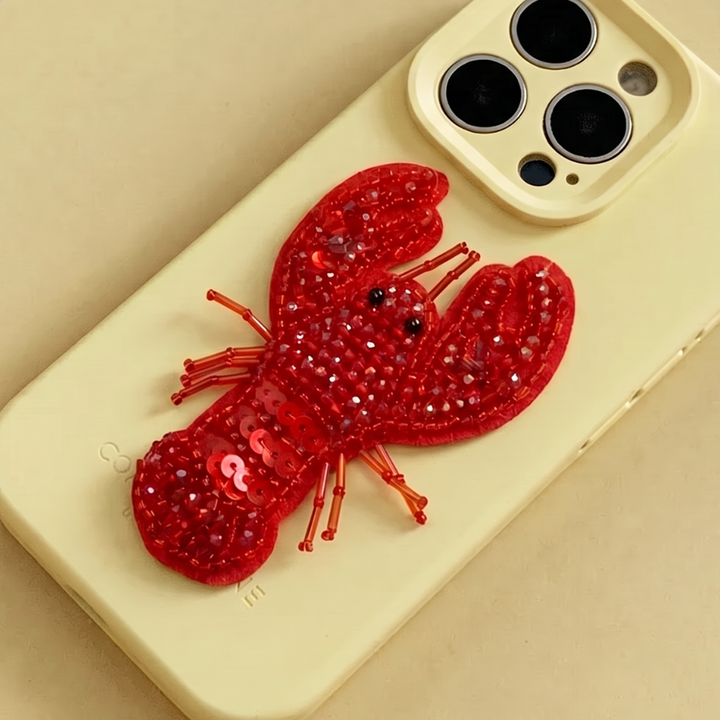 Red 3D Lobster made from beads and sequins, on top of a yellow Coconut Lane Phone Case. 