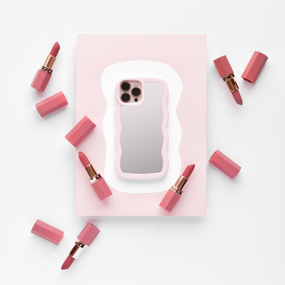 A phone with the Curvy Mirror Phone Case in pink from Coconut Lane lies on a pink and white background, surrounded by eight pink lipstick tubes, some with their caps off, revealing the lipsticks. The composition is minimalistic with a soft color palette.