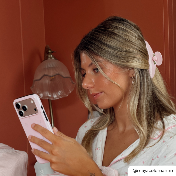 A woman with blonde-highlighted hair wearing a pink hair claw clip and a white pajama top with pink embroidery, holding a light pink smartphone in a bedroom setting with a terracotta wall and a vintage-style lamp.