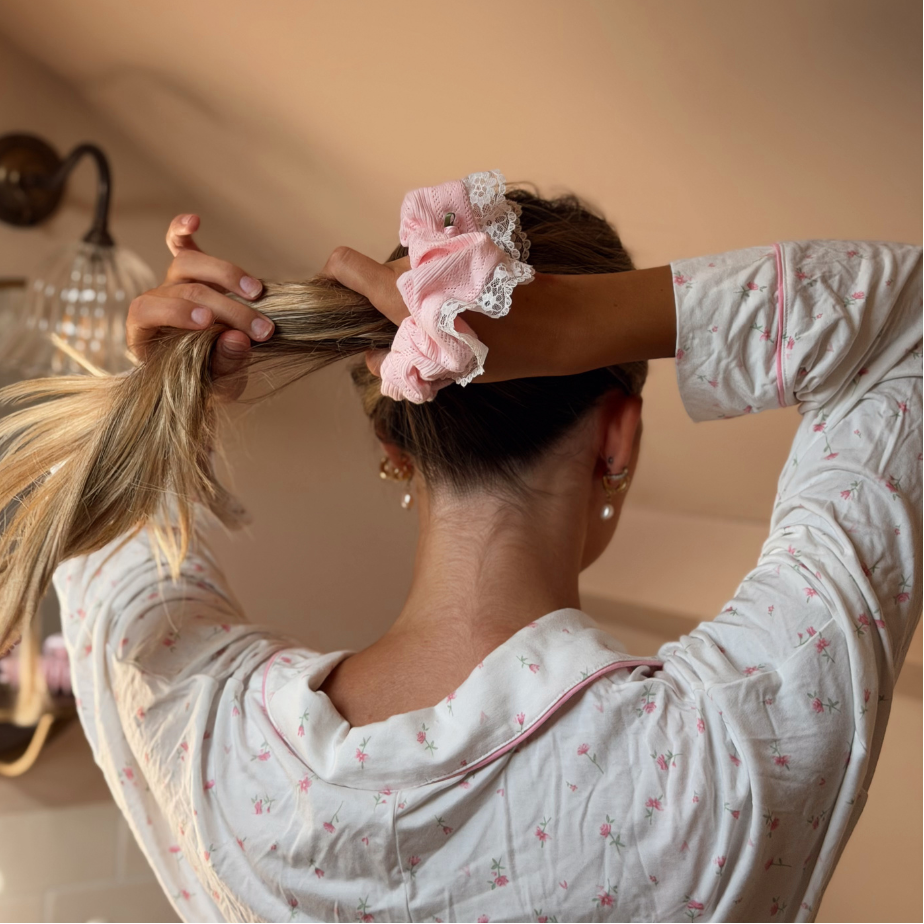 A woman with long blonde-highlighted hair seen from behind, gathering her hair with one hand while wearing a light pink scrunchie with white lace trim. She is wearing a white long-sleeve pajama top with a small pink floral print in a warm-toned room.