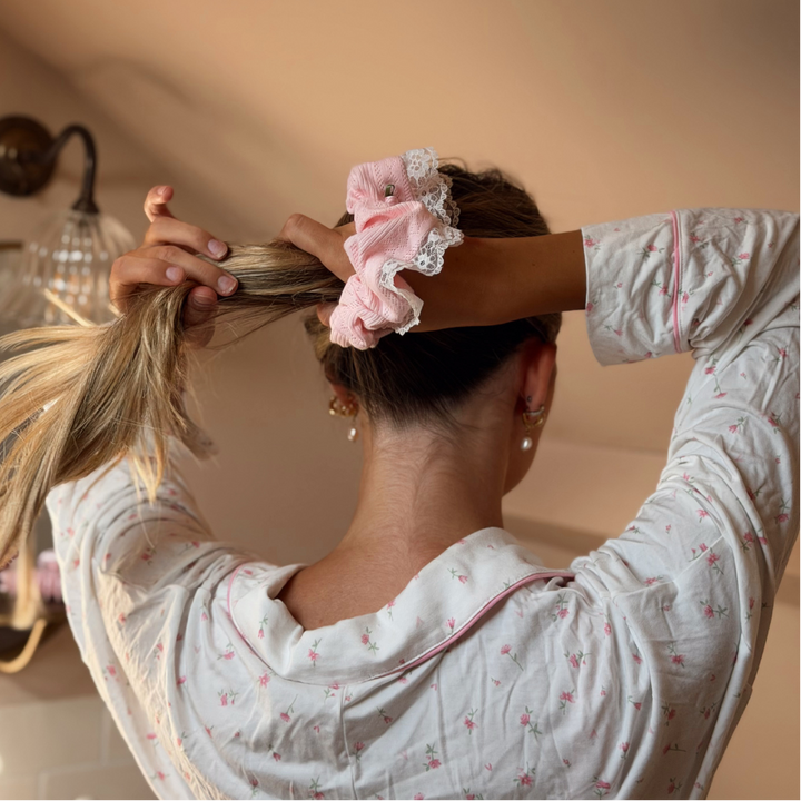 A woman with long blonde-highlighted hair seen from behind, gathering her hair with one hand while wearing a light pink scrunchie with white lace trim. She is wearing a white long-sleeve pajama top with a small pink floral print in a warm-toned room.