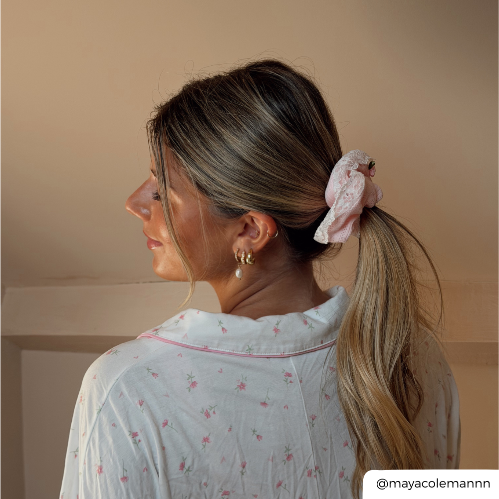 A woman shown from the back and side profile wearing a light pink floral scrunchie in her ponytail, paired with a white floral pajama top. The background is a solid warm beige wall.