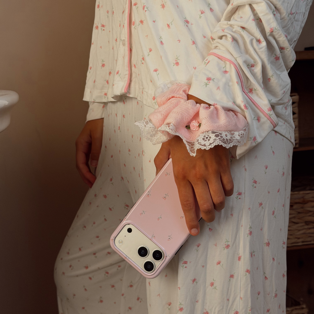 A person wearing a white long-sleeve pajama set with a small pink floral print and pink piping, holding a light pink phone case. She has a pink ruffled baby pink scrunchie on her wrist with white lace details and a small pink embroidered rose. 