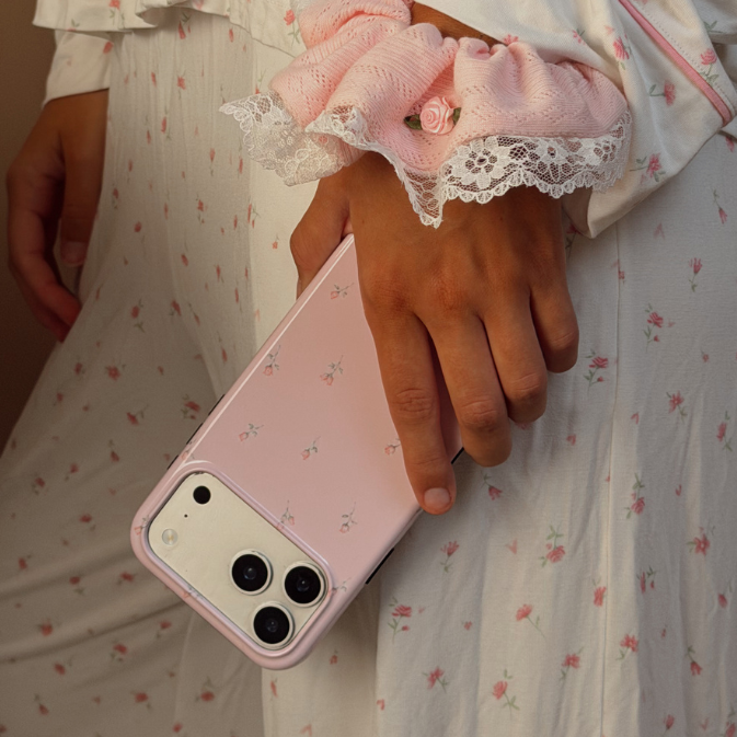 A person wearing a white floral pyjama set holds a light pink iphone case in one hand. The pyjamas have a small pink floral print. The person is wearing a pink scrunchie on her wrist with lace detail.