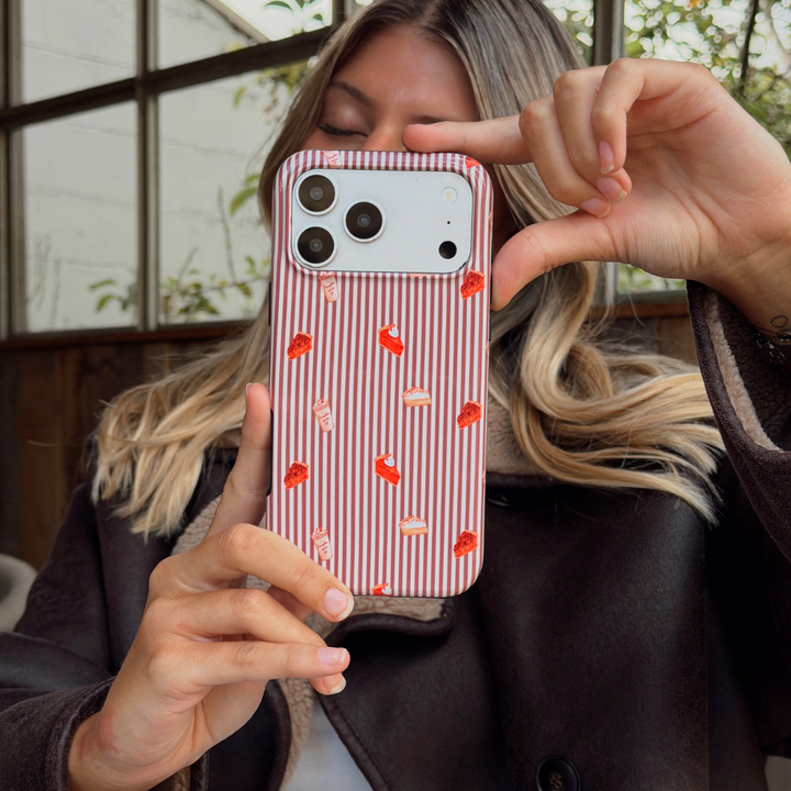 Woman wearing a brown aviator jacket holding up an iphone taking a selfie. The phone is in an autumnal case, with brown vertical stripes, and small prints of pumpkin spice lattes and sweet treats.