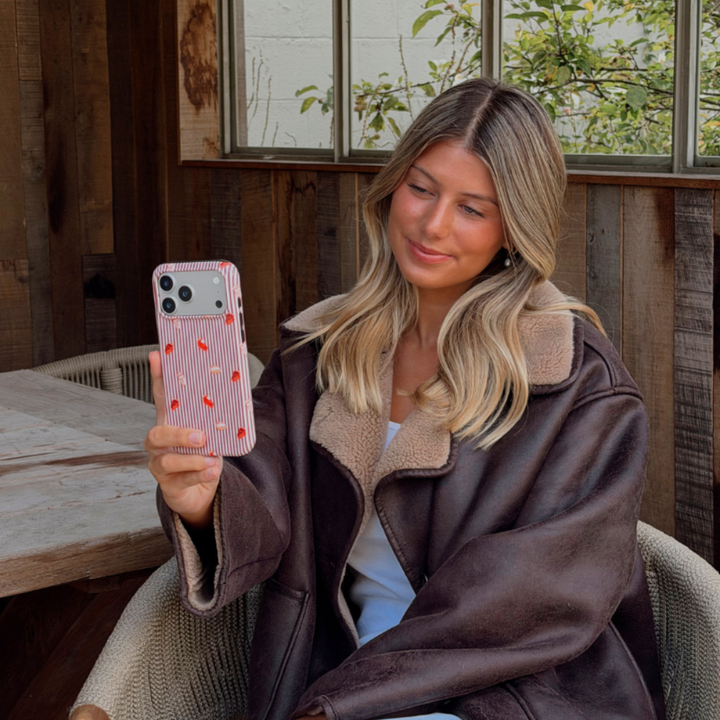 Woman wearing a brown aviator jacket holding up an iphone taking a selfie. The phone is in an autumnal case, with brown vertical stripes, and small prints of pumpkin spice lattes and sweet treats.