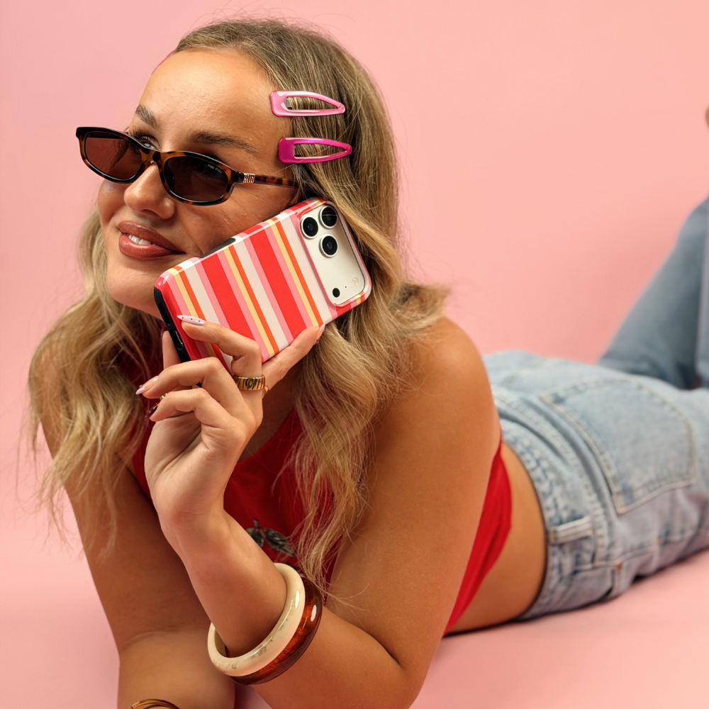 A woman lying on a pink surface wearing cat-eye sunglasses, pink hair clips, a red top, and denim shorts while holding a striped hot pink, orange, yellow, and white.