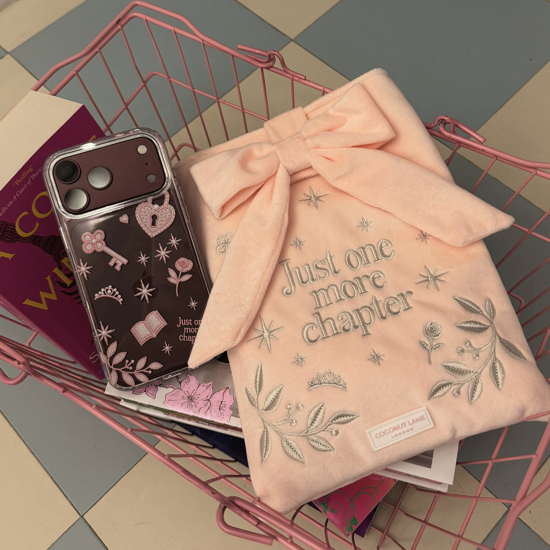 Romance Girly Clear Phone Case showing intricate pink key, heart padlock, floral and star details, placed in a pink shopping basket along side some books. 

Next to the phone is the matching, soft blush pink velvet with grey embroidered floral details, stars and 'Just One More Chapter' lettering, finished with a large pink velvet bow and Coconut Lane logo