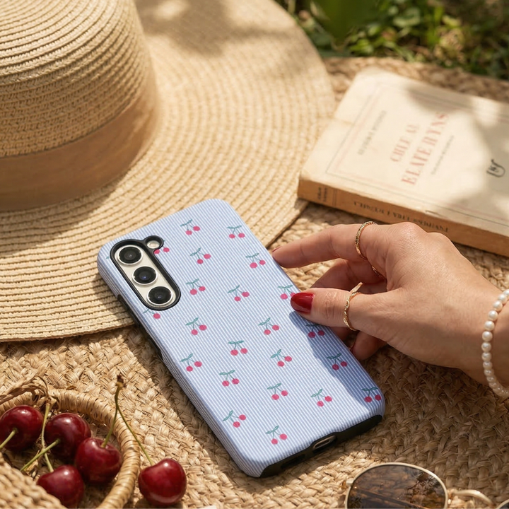 Phone case with cherry pattern held by a hand, surrounded by a straw hat, book, and cherries on a woven surface.