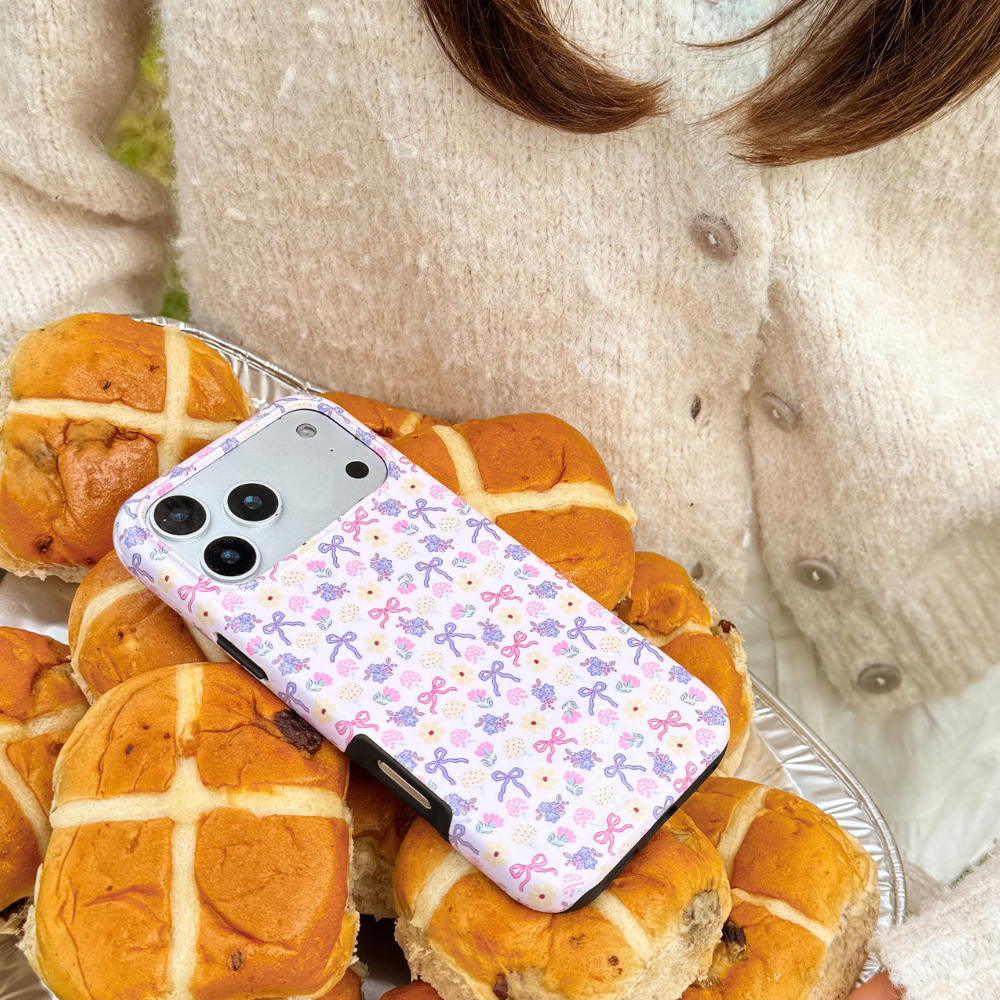 Girl in cream cardigan holding her purple phone case with bows and flowers on it called spring bows tough magsafe phone case on top of hot cross buns