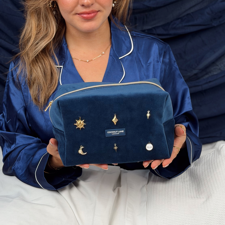 Woman wearing satin navy blue pyjamas holding a Stardust After Dark makeup bag. Dark navy blue velvet material with gold zip, and sewn-on gold celestial-themed charms including a sun, moon, star, diamond shape and 11:11 charm.  
