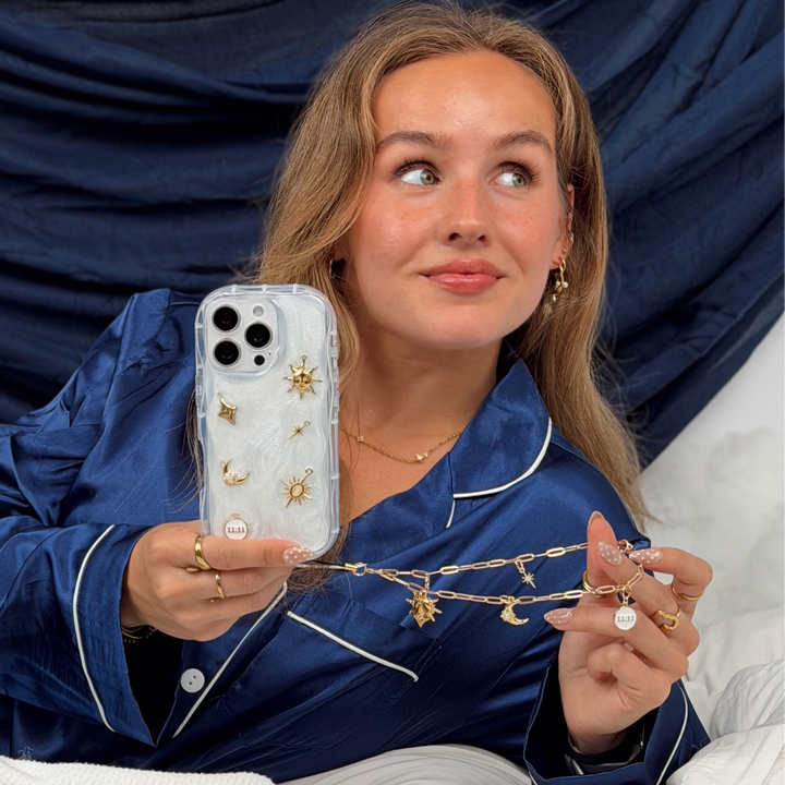 Woman wearing satin navy pyjamas laying in a bed with white bedding, She is holding a Stardust after dark charm phone case. A pearl base with luxe gold celestial charms including a moon, sun, star, diamond shape and 11:11 charm. 
