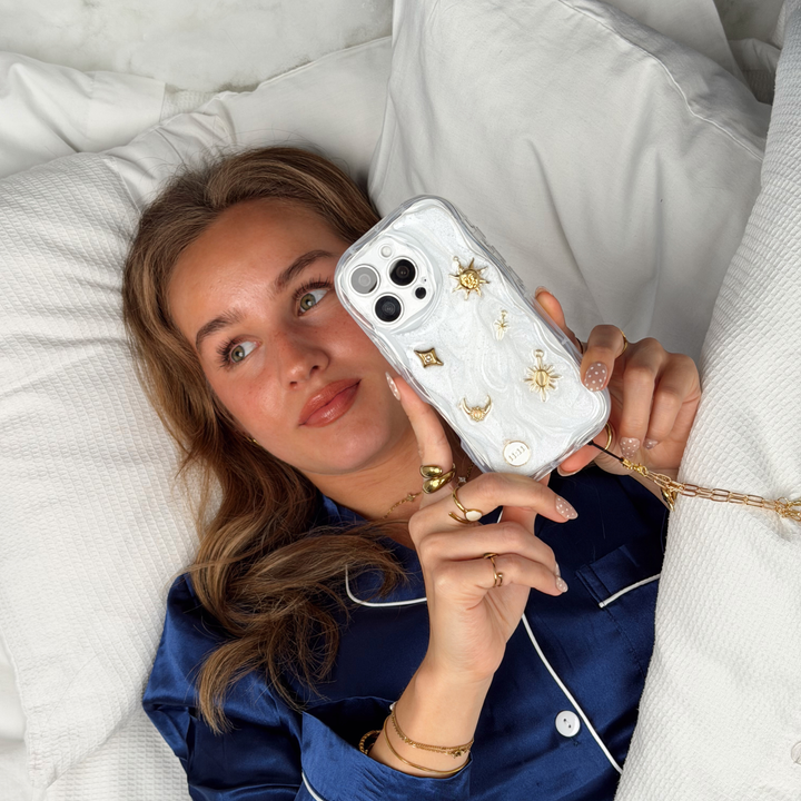 Woman wearing satin navy pyjamas laying in a bed with white bedding, She is holding a Stardust after dark charm phone case. A pearl base with luxe gold celestial charms including a moon, sun, star, diamond shape and 11:11 charm. 