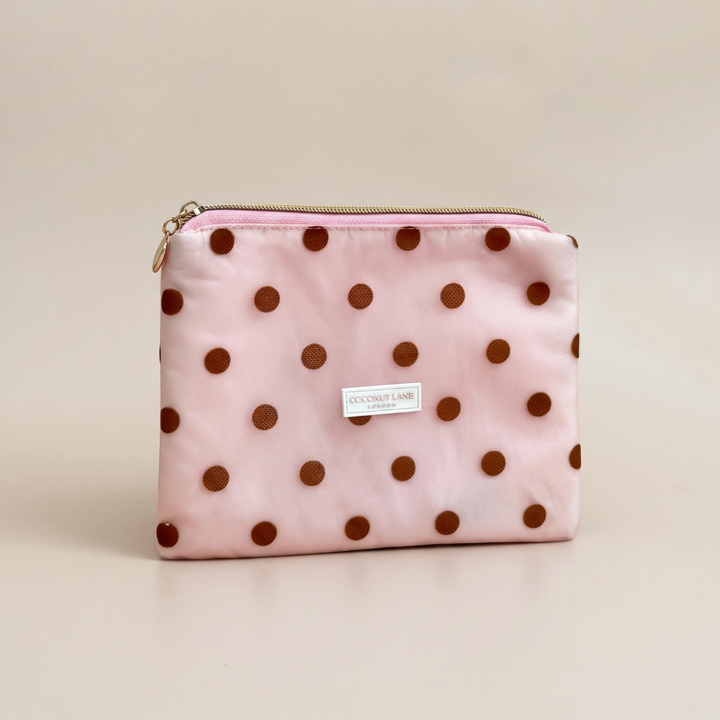 Pink mesh pouch with brown polka dots, a gold top zip with a heart-shaped zip pull, and a white Coconut Lane London logo badge on the front, shown on a beige background.