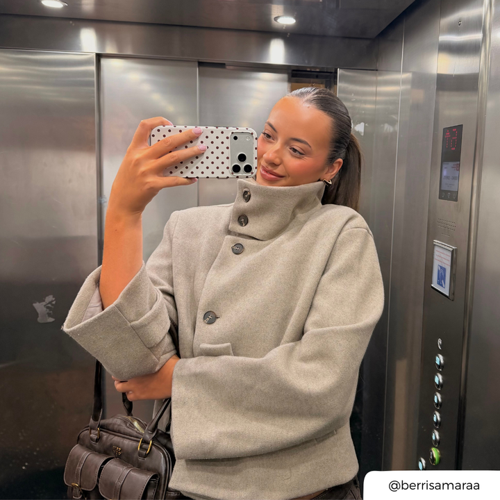 Woman with brunette hair tied in a ponytail holds up an iphone taking a mirror selfie in a lift. The phone is in a cream and brown polka dot phone case. 