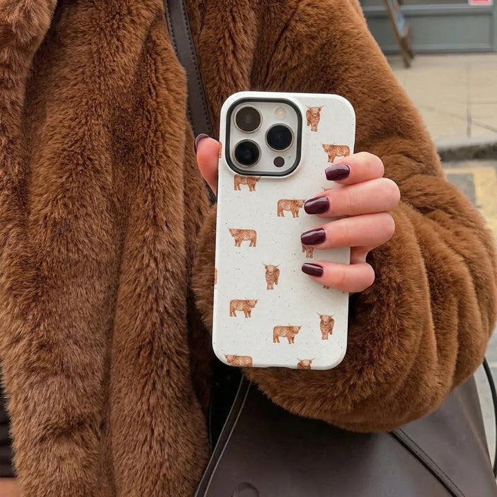 Hand with dark nails holds a Coconut Lane Highland Cow phone case; brown faux fur coat and black bag also visible.