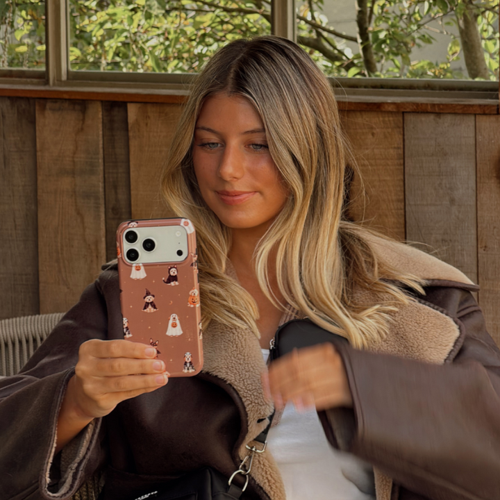 Woman in a brown aviator jacket, holding a phone case with dogs wearing spooky outfits on a brown background.