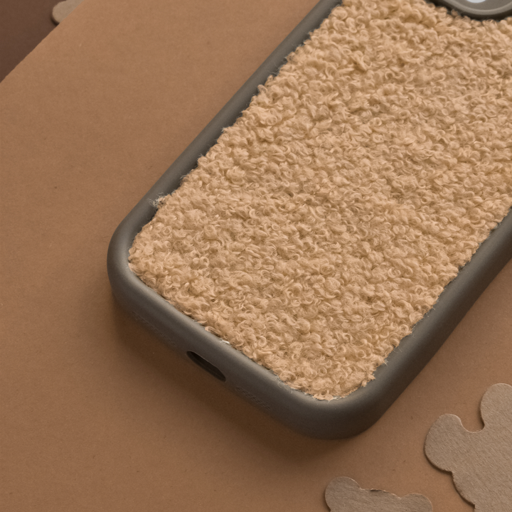 A smartphone with a Cosy Teddy Phone Case - Salted Caramel by Coconut Lane is placed on a brown surface. The case features an extra protective, beige, textured teddy material with a rough, fuzzy appearance and a dark gray border. Several abstract cardboard cutouts are scattered around it.