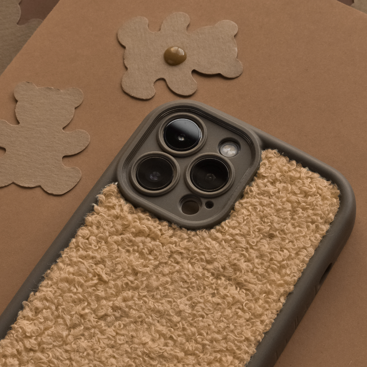 A close-up of a smartphone with Coconut Lane's Cosy Teddy Phone Case in Salted Caramel, featuring three prominent camera lenses. The phone is placed on a brown surface with cut-out teddy bear shapes in the background, matching the earthy tones of this extra protective case.