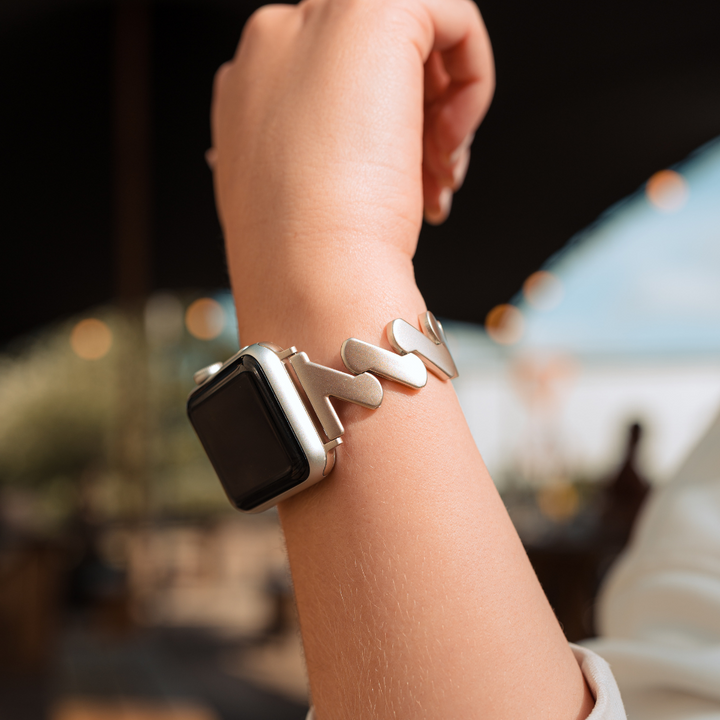 A person wearing a smartwatch adorned with a sleek, silver, and stylish Get Wavy Apple Watch Strap in Starlight by Coconut Lane. The smartwatch screen is black and currently not displaying any information. The background is slightly blurred, showcasing a vibrant outdoor scene with beautiful bokeh lights. This stainless steel metal adjustable band fits ALL Apple Watch models perfectly.