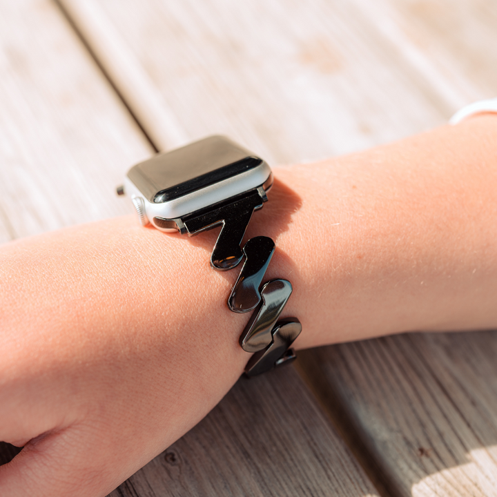 A close-up of a person's wrist wearing the Coconut Lane Get Wavy Apple Watch Strap - Black, featuring an adjustable zigzag-patterned bracelet. The hand rests on a wooden surface, highlighting the natural texture of the wooden table in the background.