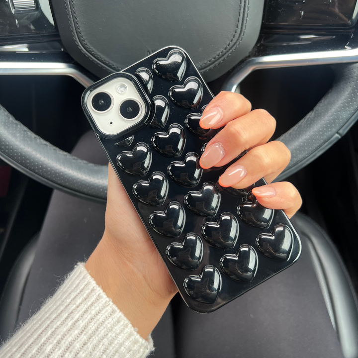 A hand with manicured nails holds an iPhone15 encased in the highly protective, Glossy Black 3D Hearts Phone Case by Coconut Lane. The background reveals a car steering wheel and part of the dashboard, while the person’s sleeve is partially covered by a light-colored sweater.