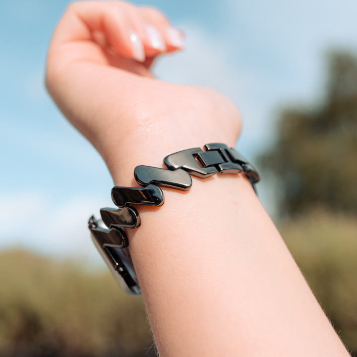 A close-up of a person's wrist wearing the Coconut Lane Get Wavy Apple Watch Strap - Black, featuring a black, metallic, rectangular-link design. The background is slightly blurred, showcasing greenery and a clear blue sky. The adjustable strap perfectly complements the Apple Watch. The person's hand is raised with only part of a finger visible.