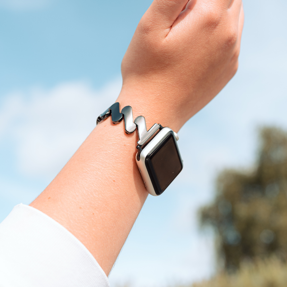 A close-up of a wrist wearing an Apple Watch adorned with Coconut Lane's Get Wavy Apple Watch Strap - Black. The background features a blurred outdoor scene with a blue sky and some greenery, while the smartwatch screen is black and appears to be off.