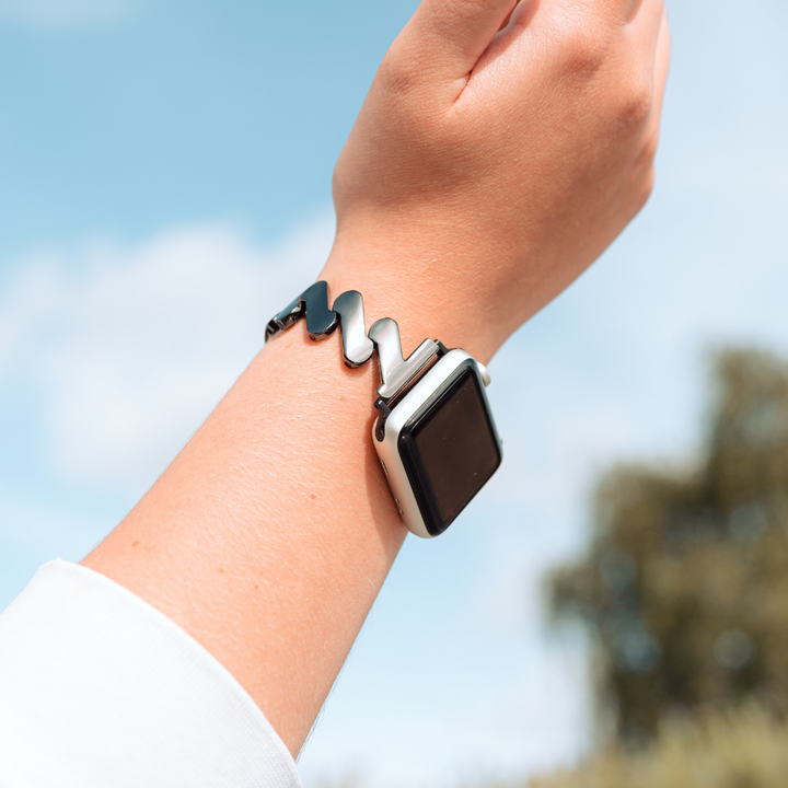 A close-up of a wrist wearing an Apple Watch adorned with Coconut Lane's Get Wavy Apple Watch Strap - Black. The background features a blurred outdoor scene with a blue sky and some greenery, while the smartwatch screen is black and appears to be off.