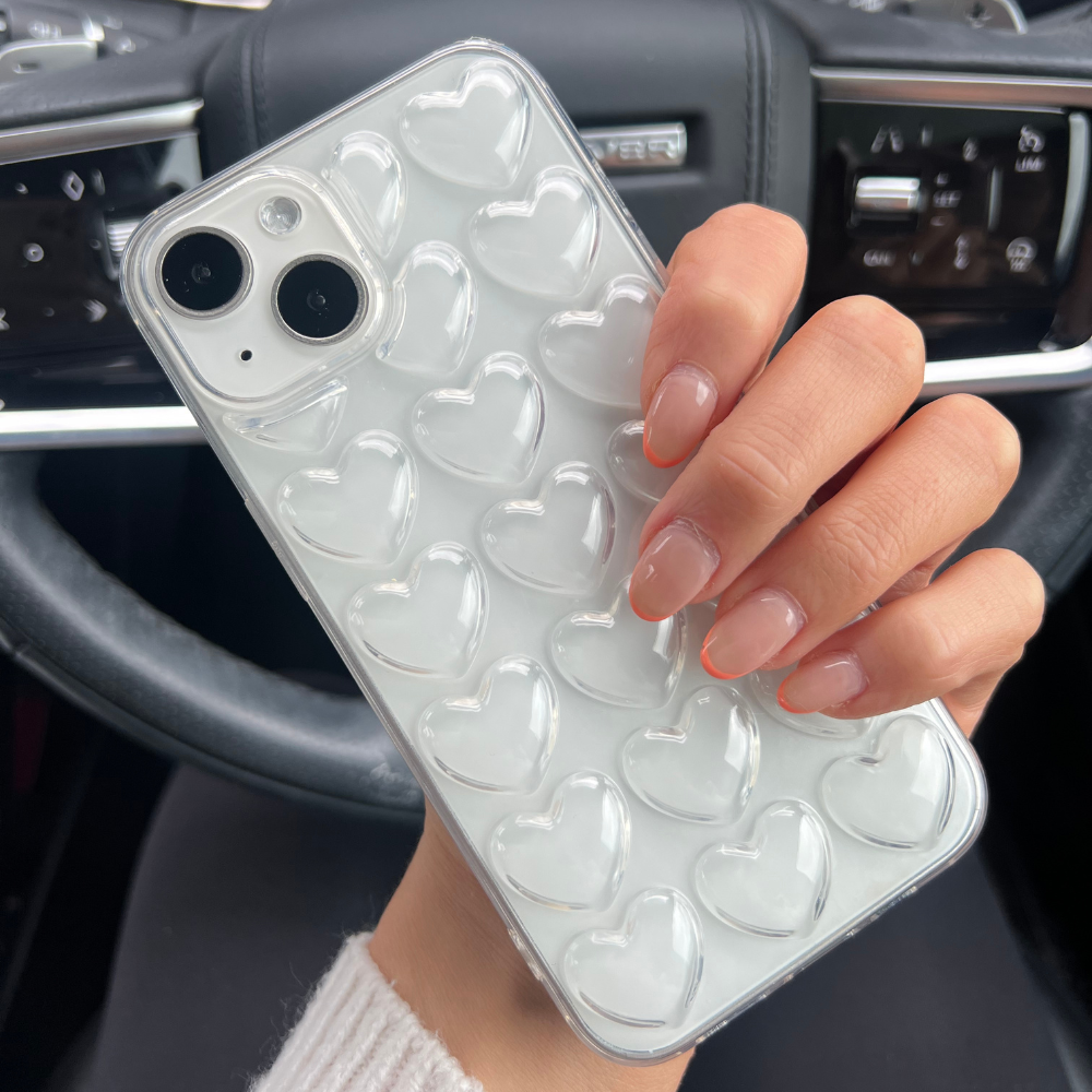 A hand with neatly manicured nails holds a Coconut Lane 3D Hearts Phone Case - Clear designed for the white iPhone15. The background reveals the interior of a car, highlighting the steering wheel and dashboard controls.