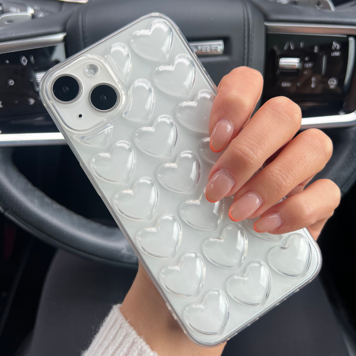 A hand with neatly manicured nails holds a Coconut Lane 3D Hearts Phone Case - Clear designed for the white iPhone15. The background reveals the interior of a car, highlighting the steering wheel and dashboard controls.