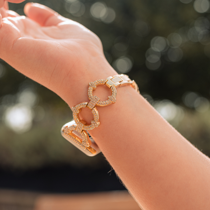 A wrist adorned with a Coconut Lane Diamante Bracelet Apple Watch Strap featuring three interconnected geometric shapes, each encrusted with sparkling stones. The skin has a warm tone, and the background is blurred greenery, suggesting an outdoor setting in natural light. It perfectly complements the elegant design of the bracelet.