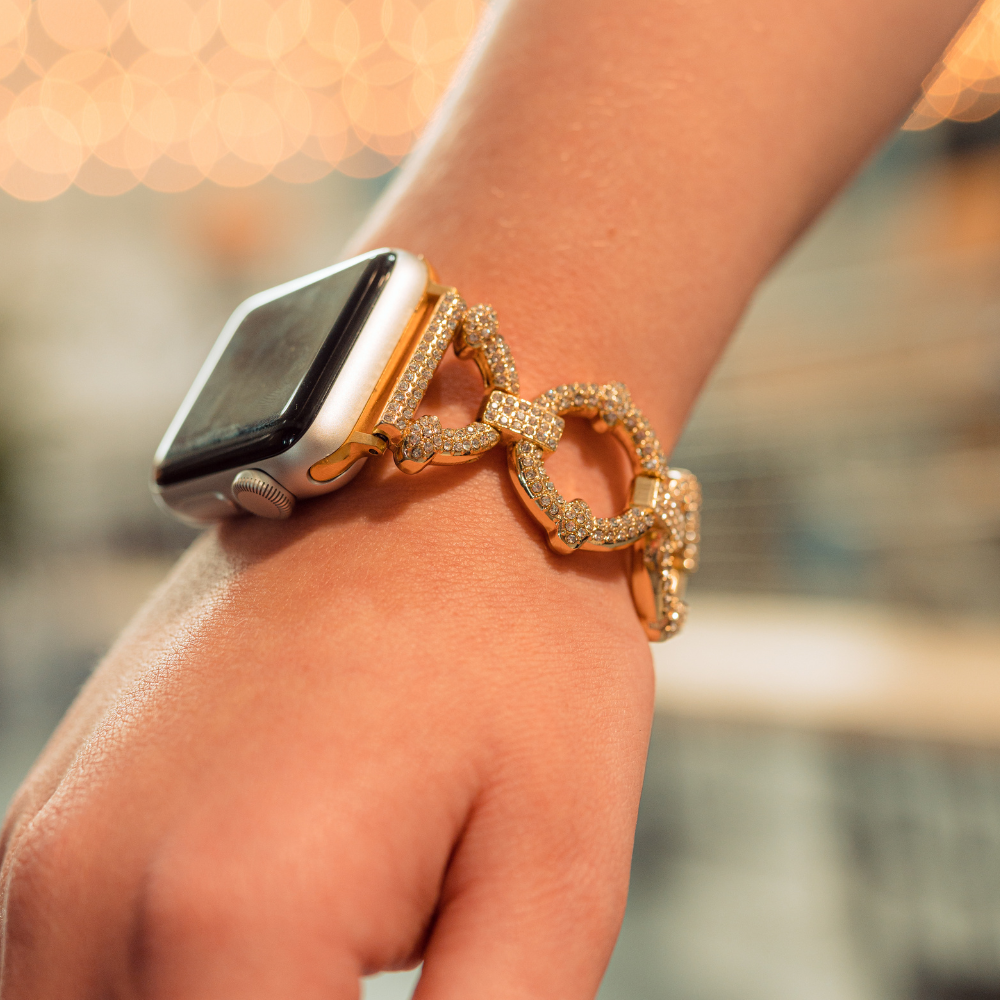 A close-up of a person's wrist wearing an Apple Watch with the distinctive Coconut Lane Diamante Bracelet Apple Watch Strap, adorned with sparkling gemstones. The background is softly blurred with warm, bokeh lights, drawing focus to the elegant accessory.