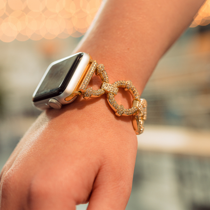 A close-up of a person's wrist wearing an Apple Watch with the distinctive Coconut Lane Diamante Bracelet Apple Watch Strap, adorned with sparkling gemstones. The background is softly blurred with warm, bokeh lights, drawing focus to the elegant accessory.