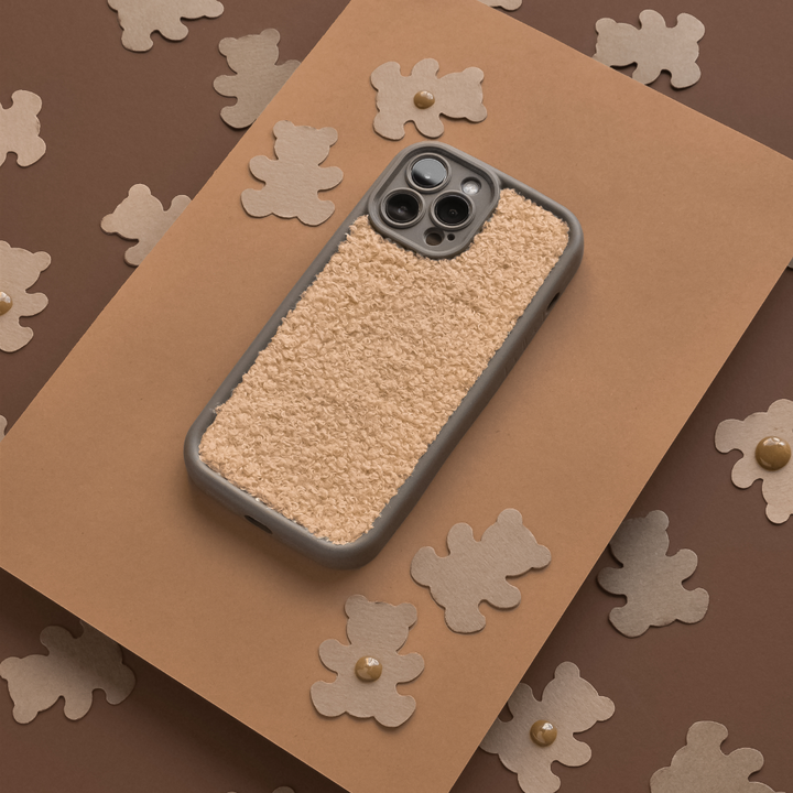 A Cosy Teddy Phone Case - Salted Caramel by Coconut Lane is placed on a brown background. The background is decorated with cut-out shapes of brown teddy bears, adding a playful and cozy vibe to the image.