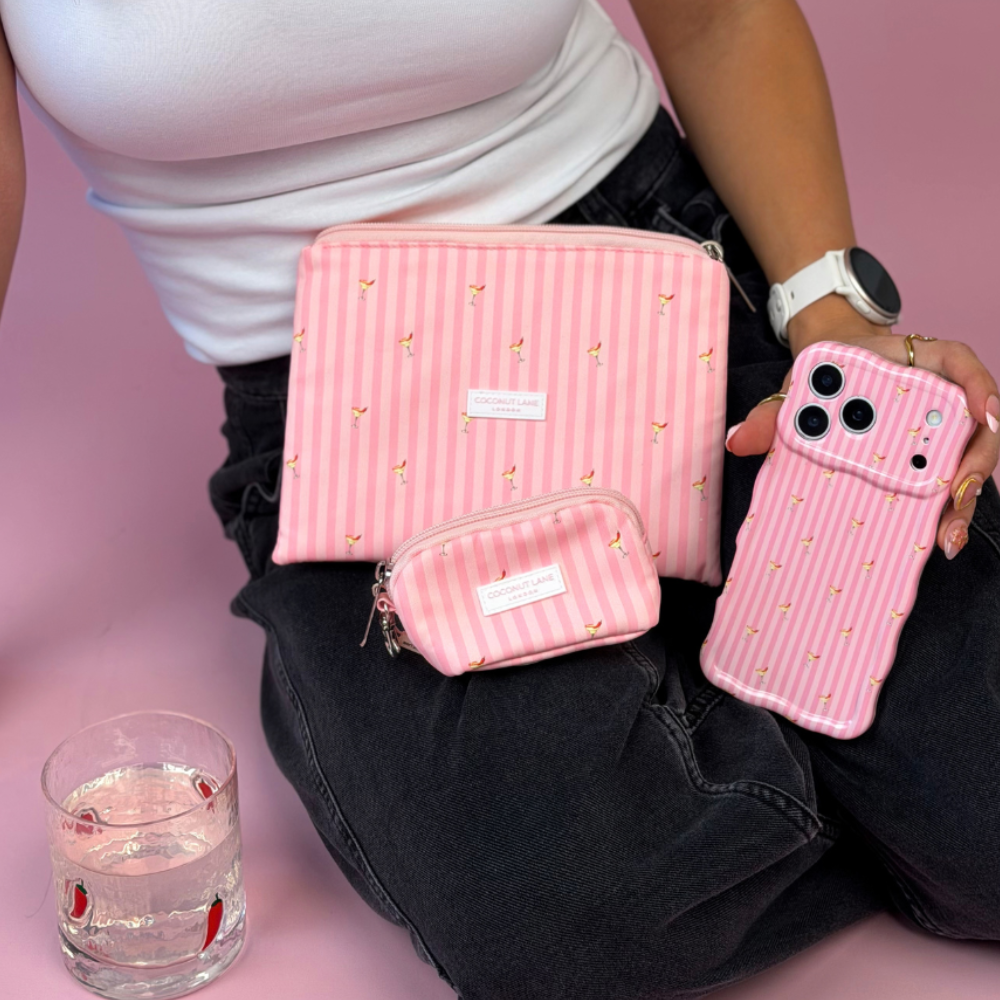 A person sitting on a pink floor wearing a white top and black jeans, holding a wavy pink striped phone case, with a pink zippered cosmetic pouch and mini keyring bag, with vertical pink stripes and small spicy margarita illustration prints.