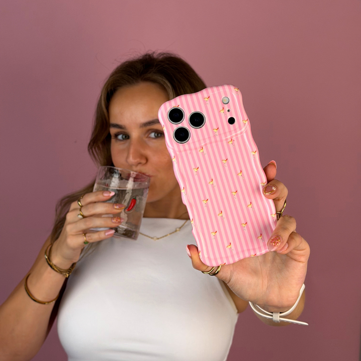 A woman taking a selfie, holding a wavy pink striped phone case with small spicy margarita illustrations, held by a person wearing a white top and black jeans against a solid pink background. She is sipping from a glass with red chillis on it. 
