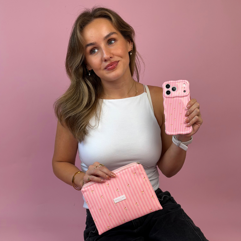 A woman wearing a white tank top and black pants holds a pink striped pouch and a pink striped wavy phone case. The pouch and phone case are decorated with small spicy margarita illustration prints.
