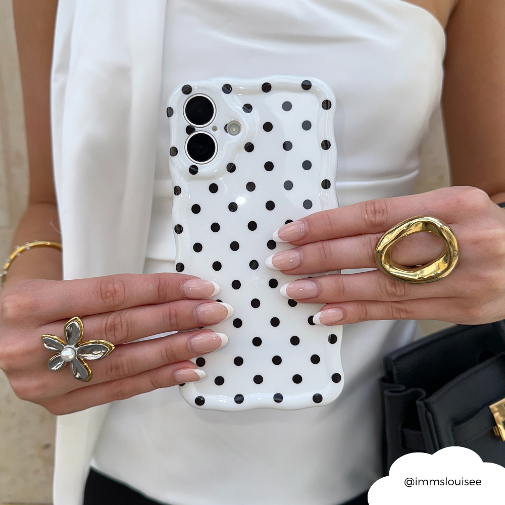 Hand with manicured nails and gold rings holds a white phone case with wavy edges and black polka dots.