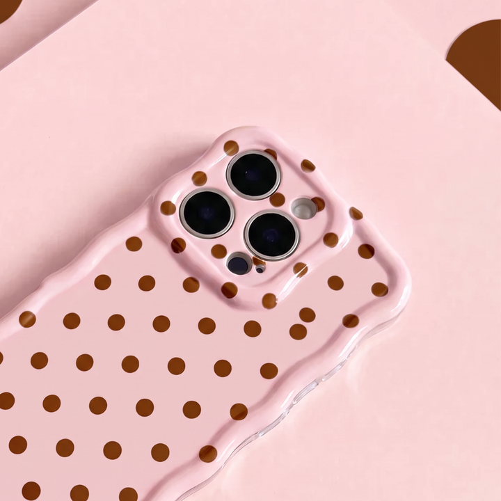 Close- up of pink Wavy Phone Case featuring a glossy blush pink finish with brown polka dots, scalloped edges and triple camera cutouts, displayed on a matching pink background.